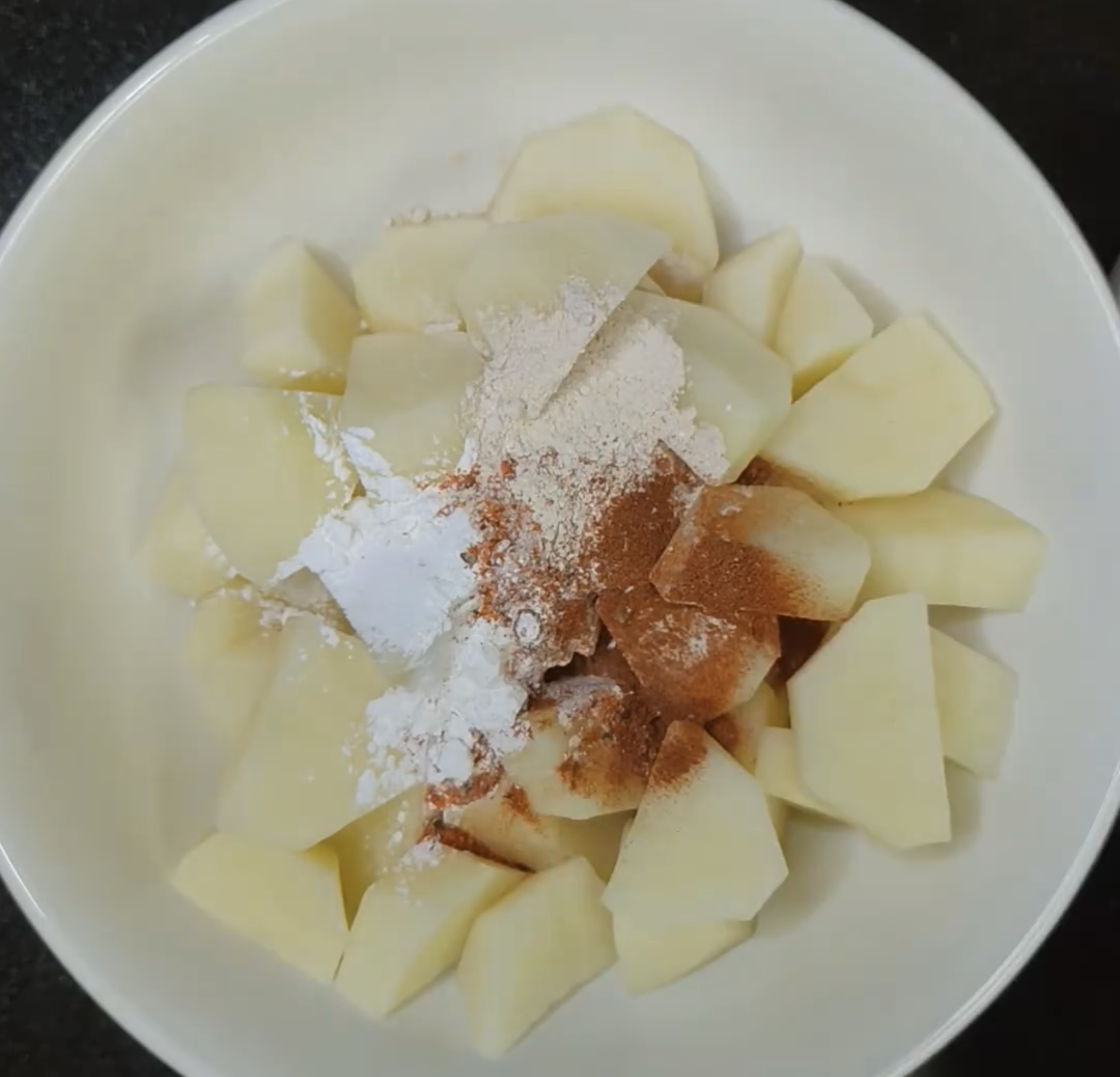 Peeled, cubed potatoes in a white mixing bowl topped with cornstarch, paprika and other seasonings