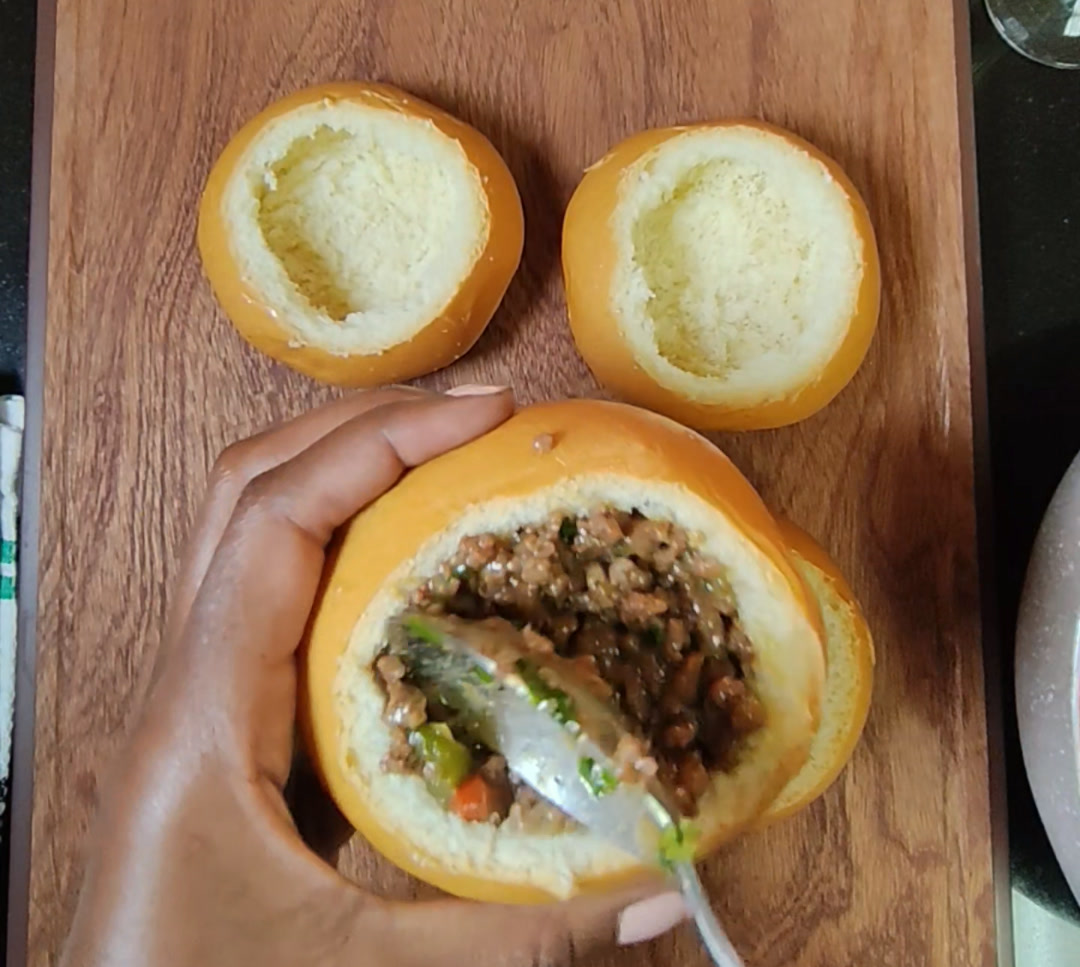 Two hollowed brioche buns on a wooden board while a hand spoons minced beef filling into a third bun