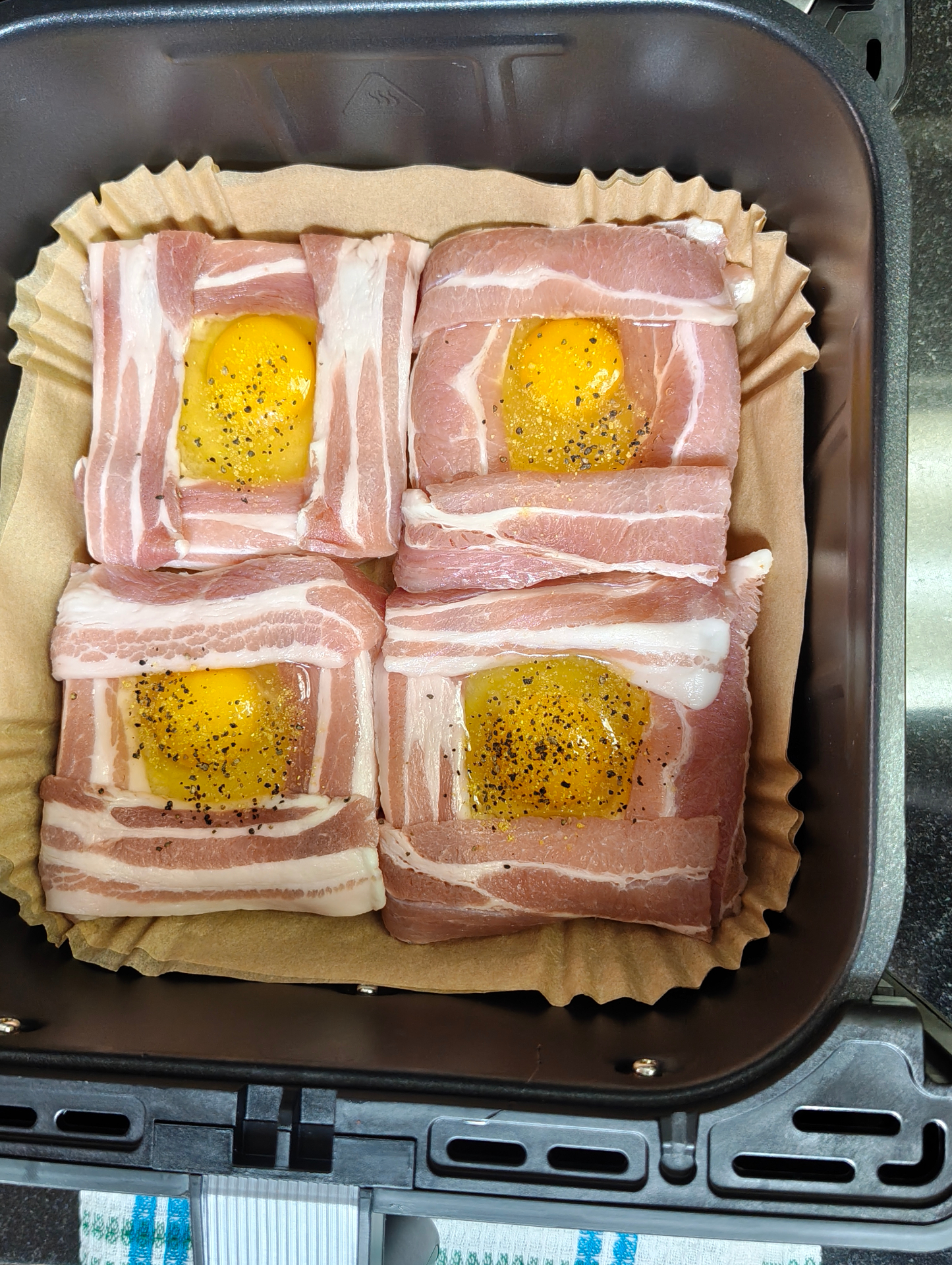 Four raw bacon-wrapped egg parcels with visible yolks arranged on a paper liner in an air fryer basket