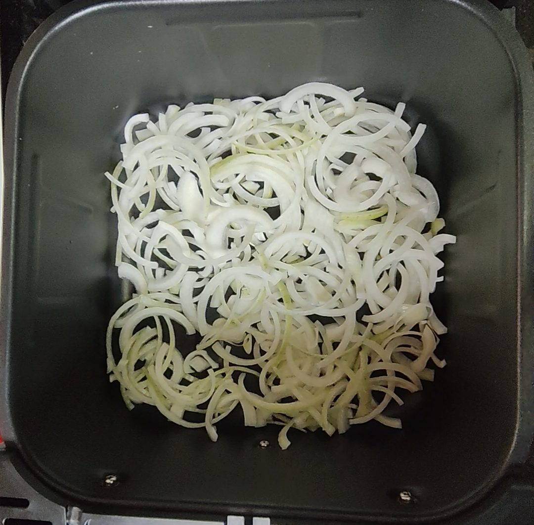Pale raw sliced onions spread across an air fryer basket before cooking