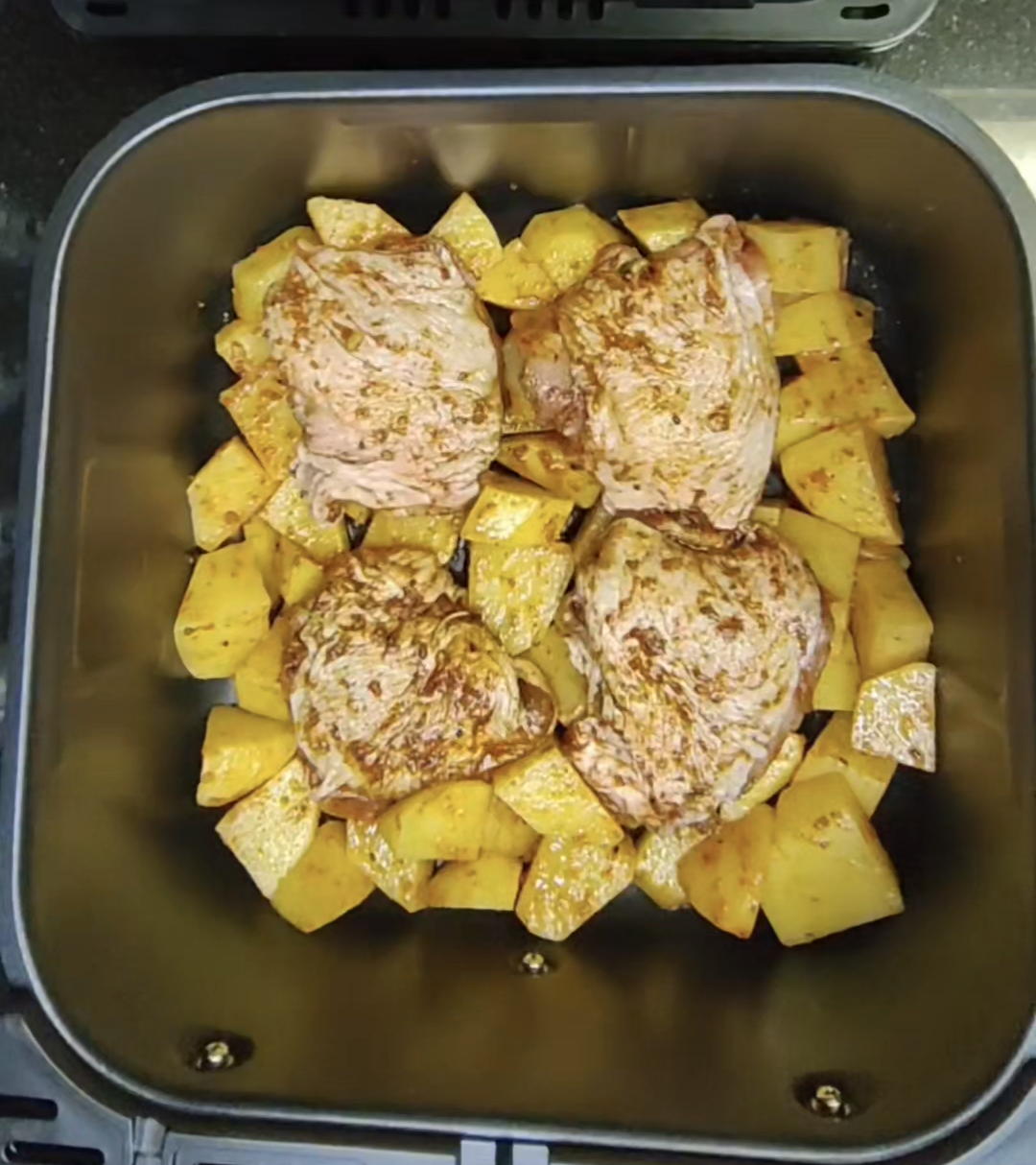 Seasoned raw chicken thighs and chunky potatoes in an air fryer basket before cooking