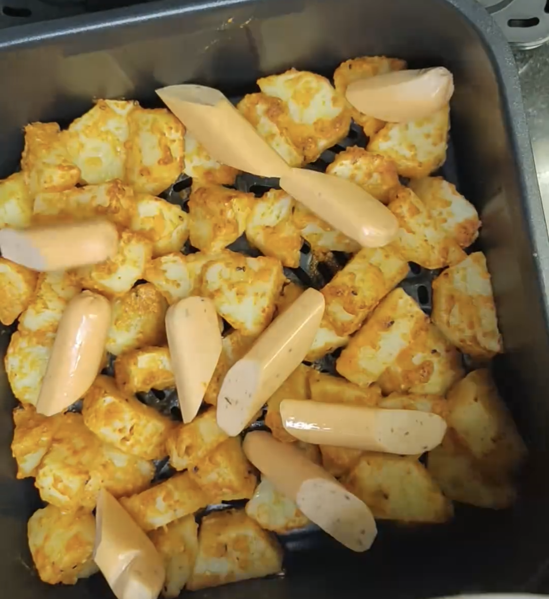 Sliced raw chicken sausages added on top of the part-cooked potatoes in the air fryer basket