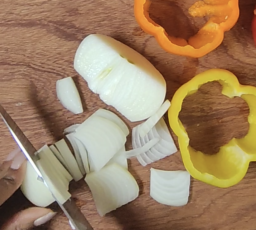 White onion being sliced into half moons with a knife on a wooden chopping board, with yellow and orange bell pepper rings nearby