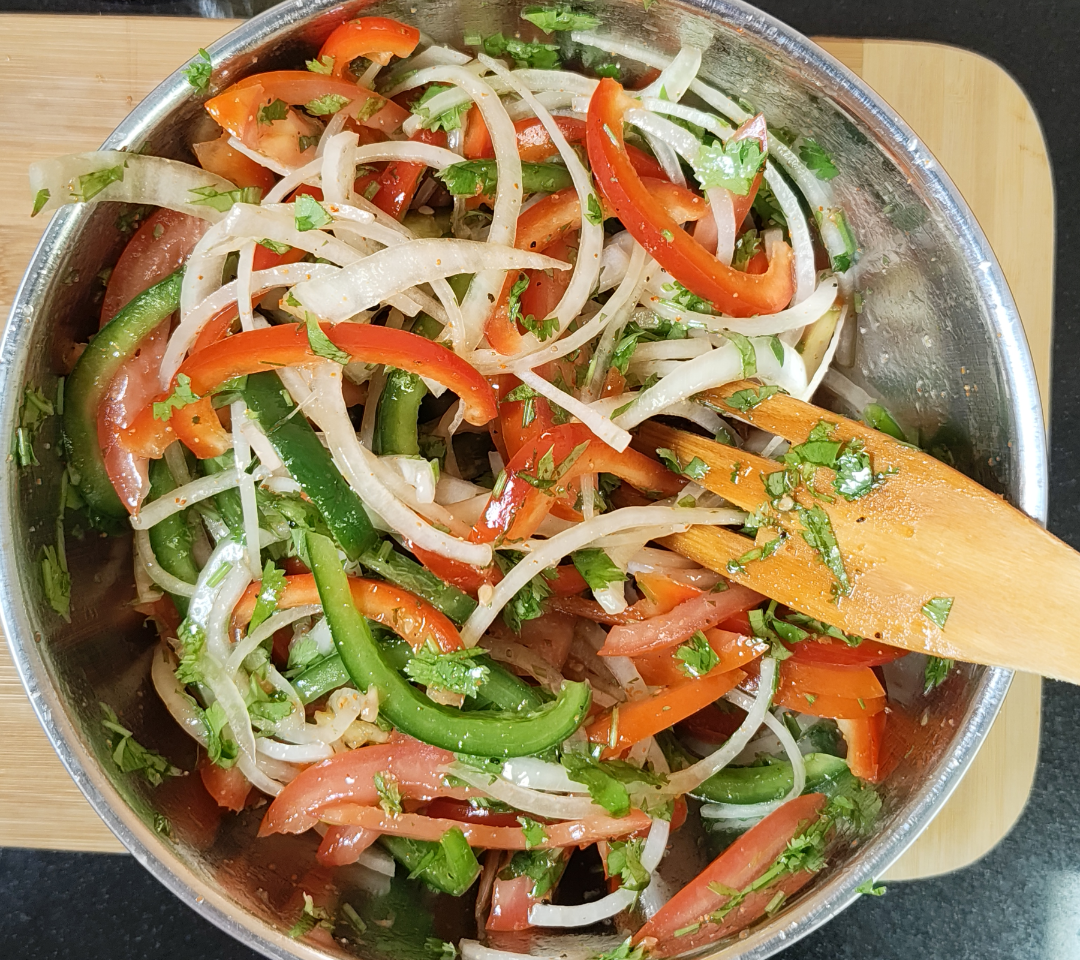 Salad in a metal mixing bowl with thinly sliced onion, tomato wedges, red and green bell pepper strips and chopped coriander
