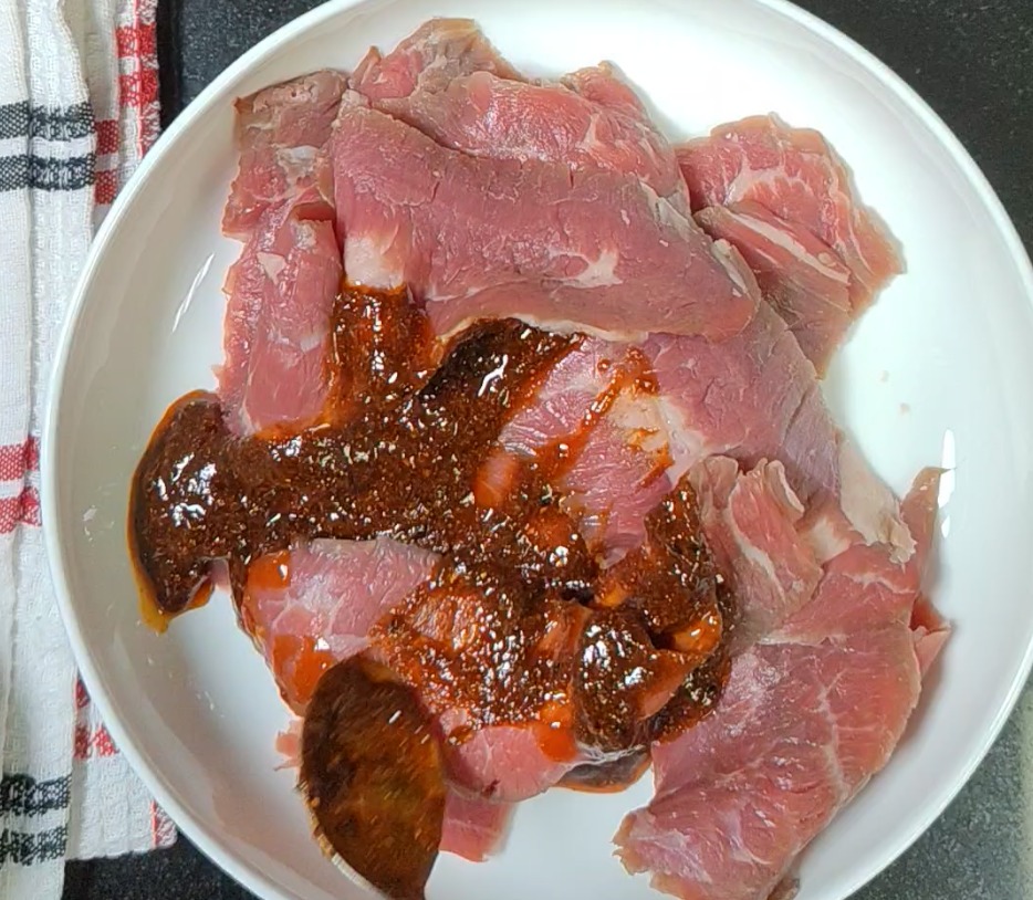 Raw skirt steak on a white plate with BBQ seasoning mixture and a wooden spoon