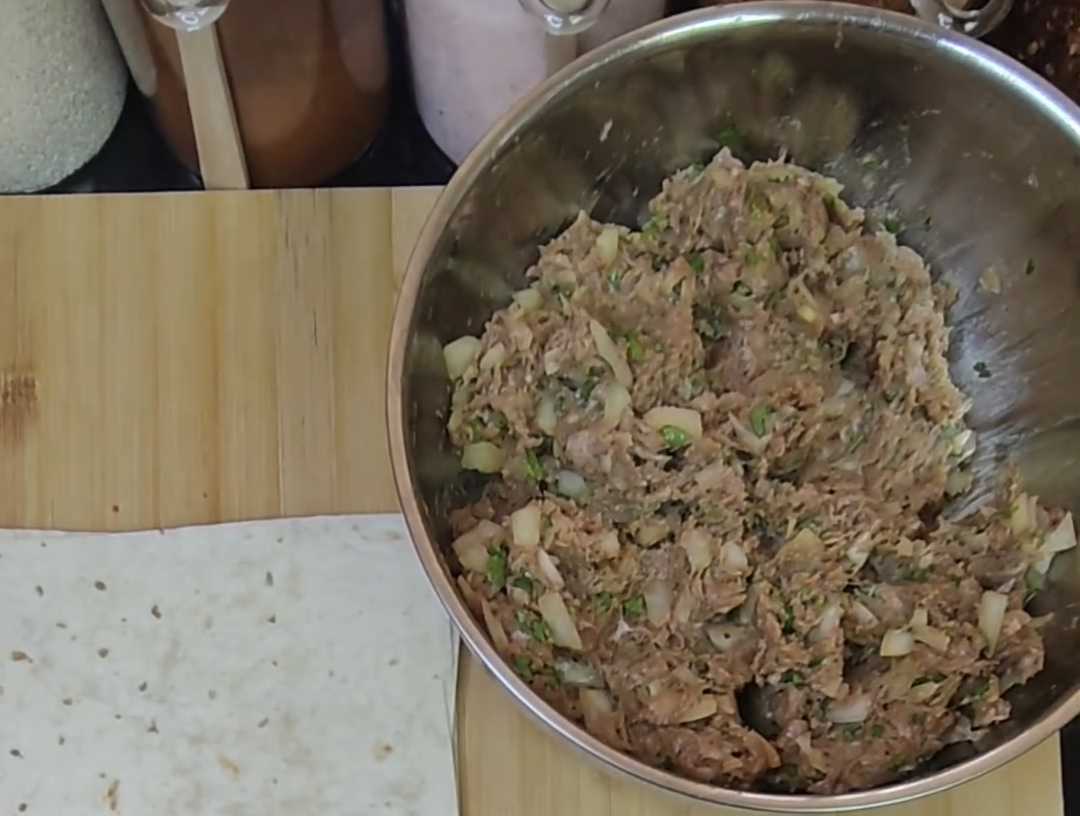Minced beef mixture with chopped onion and herbs in a stainless steel mixing bowl beside a tortilla