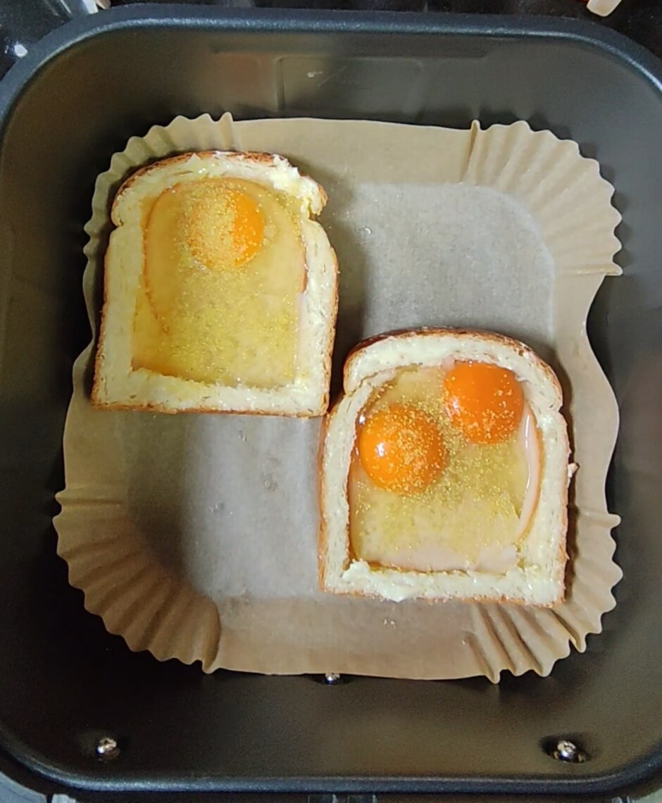 Two hollowed brioche slices in an air fryer basket on a liner, one holding one raw egg and the other two raw eggs, sprinkled with seasoning