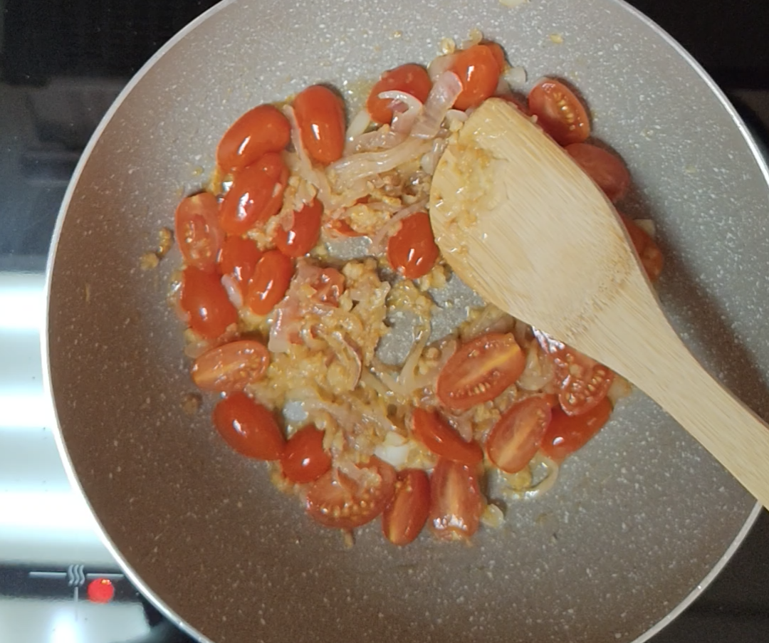 Small red tomatoes, many halved, cooking with sliced onion in a light-coloured pan with a wooden spoon