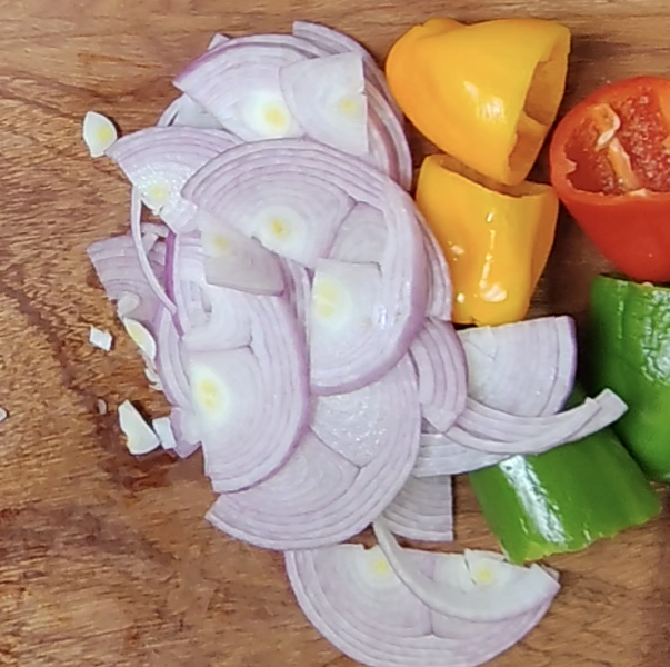 Sliced red onion beside pieces of yellow, red and green bell pepper on a wooden chopping board
