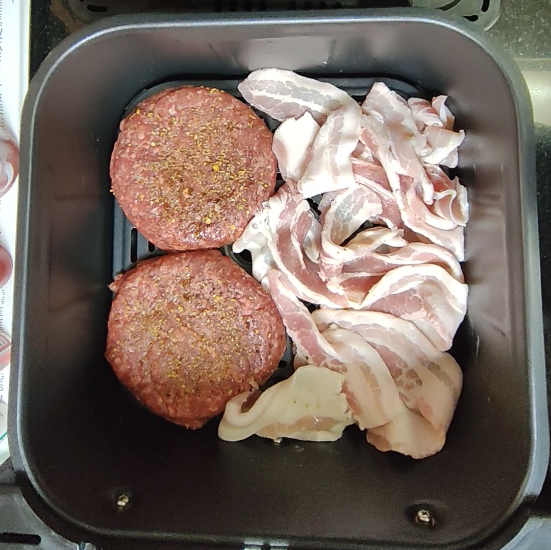 Raw burger patties and bacon in an air fryer basket before cooking