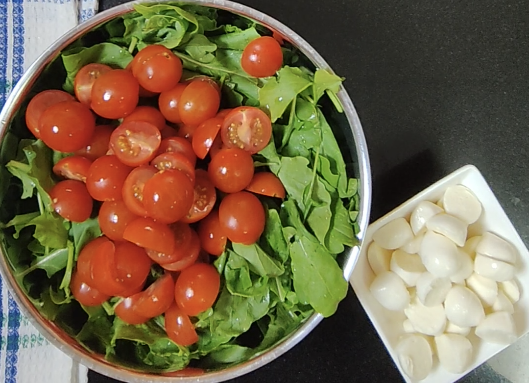 Whole and halved small red tomatoes over leafy greens in a metal bowl, with a small white dish of mozzarella beside it