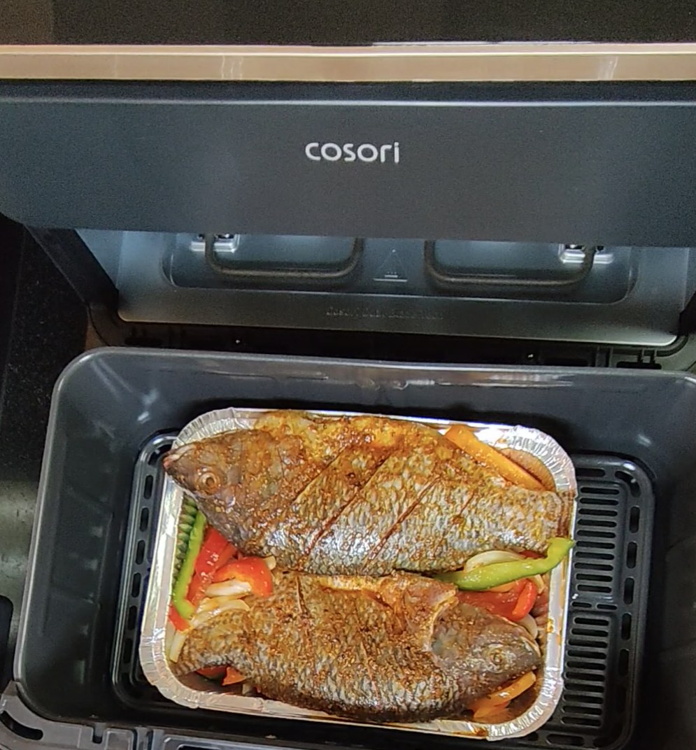 Two seasoned whole tilapia over sliced peppers and onion in an aluminium tray in an air fryer, before cooking