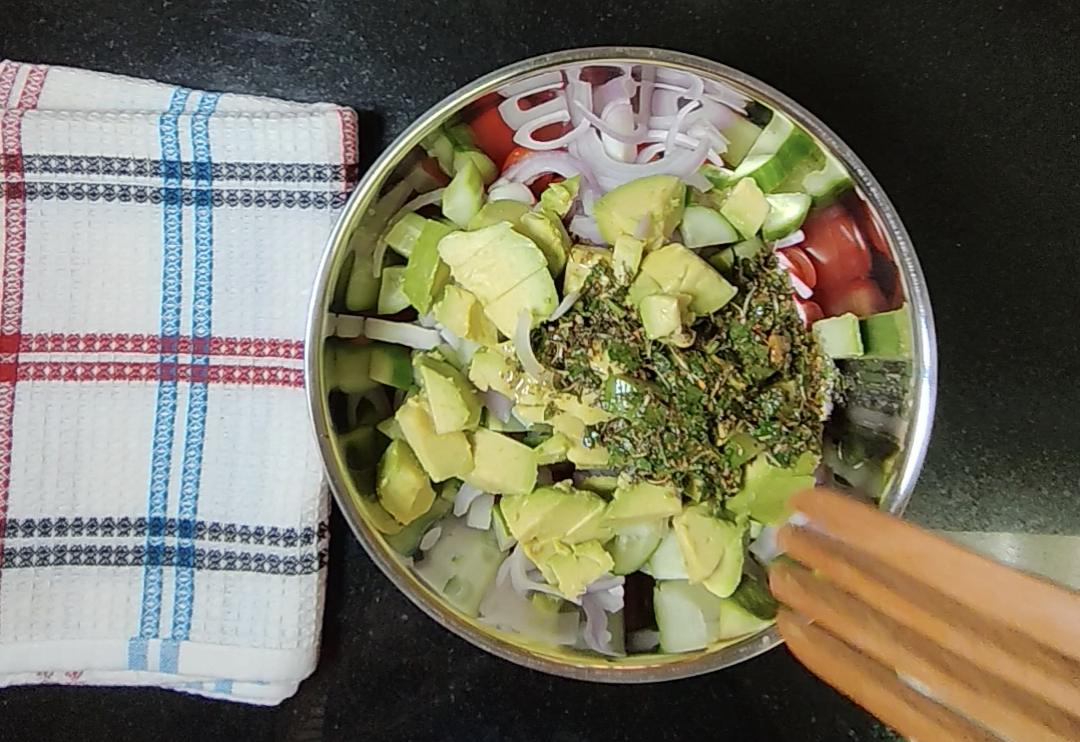 Herb and olive oil dressing with parsley added on top of the avocado, cucumber, onion and tomatoes in a metal bowl