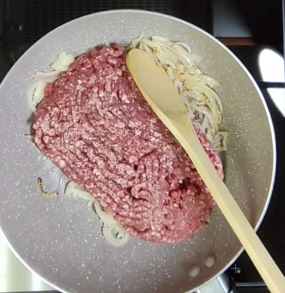 Raw minced beef added to softened sliced onion in a light-coloured pan with a wooden spoon