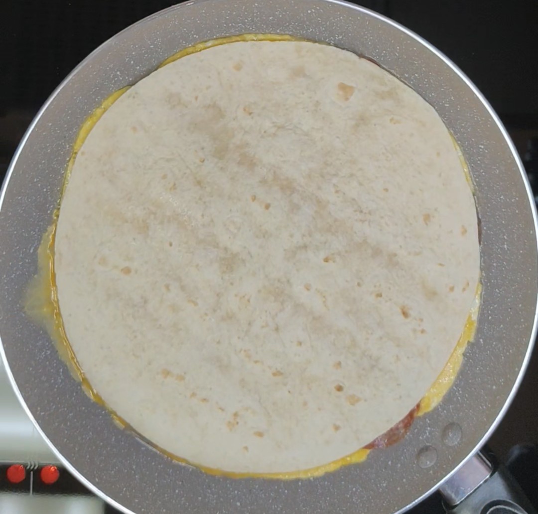 Large flour tortilla placed over eggs, salami and cheese in a frying pan