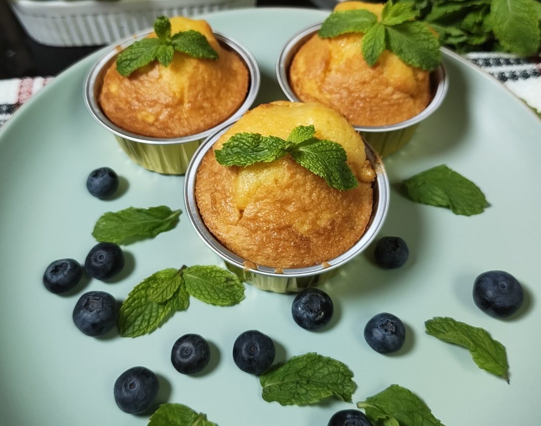 Three regular blueberry lemon muffins in disposable aluminium cups topped with mint, with fresh blueberries on a pale blue plate