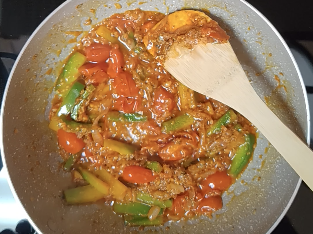 Tomatoes cooked down into a thick, saucy mixture with strips of green and yellow pepper in a light-coloured pan with a wooden spoon