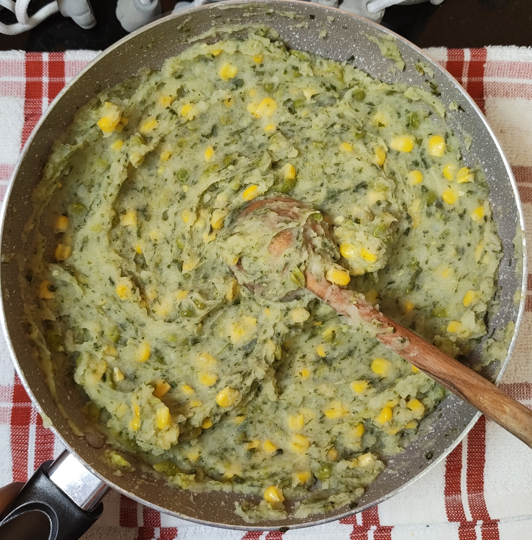 A pan of mashed mukimo with potatoes, spinach, green peas and sweet corn, with a wooden spoon resting in it