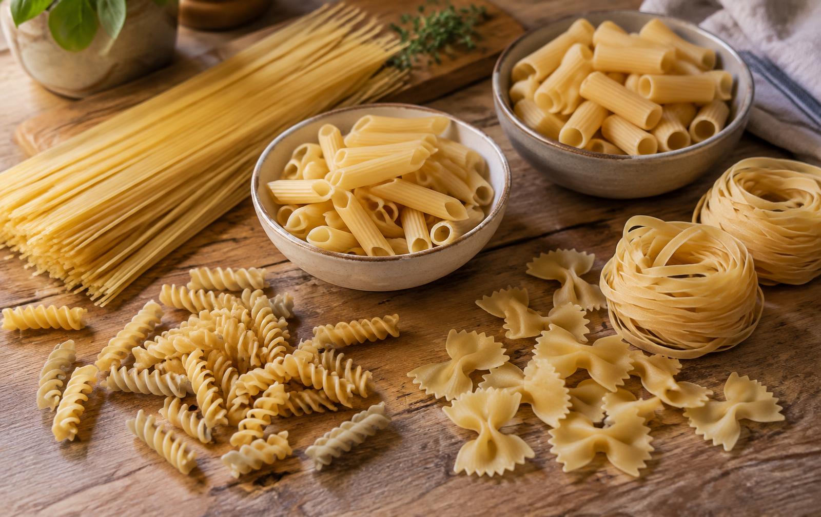 Assorted dry pasta on a wooden kitchen table, including a bundle of spaghetti, bowls of penne and rigatoni, scattered fusilli and farfalle, and nests of tagliatelle
