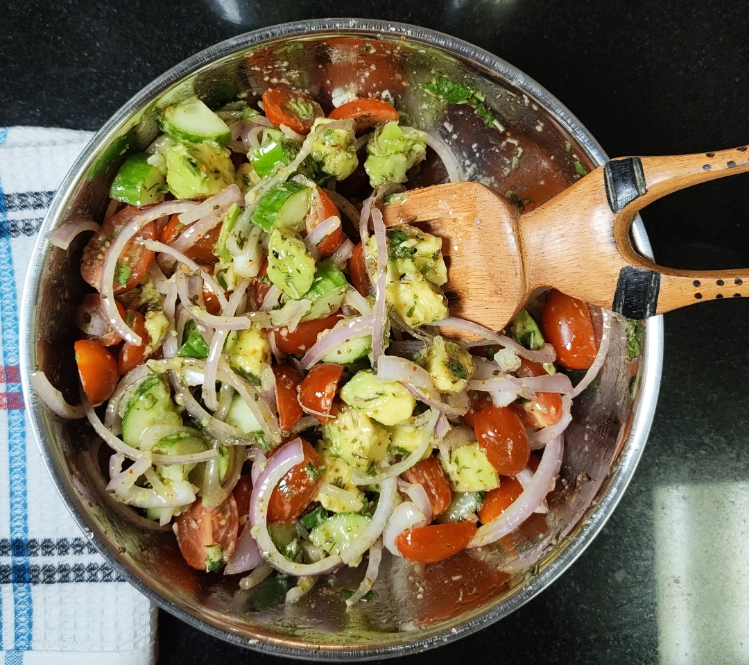 Tossed avocado, tomato and cucumber salad with sliced onion and herb dressing in a metal bowl with a wooden serving fork