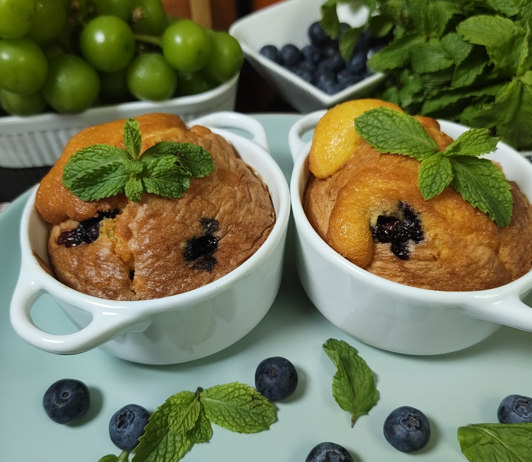 Two blueberry lemon muffins in white ramekins topped with fresh mint, with fresh blueberries on a pale blue plate